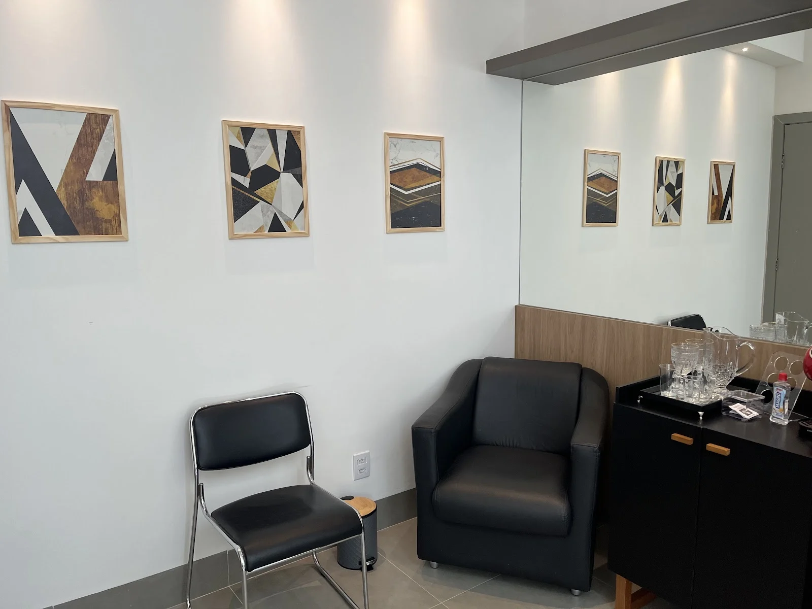 Sala de espera com poltrona e cadeira pretas, parede branca com três quadros geométricos e um aparador com copos e jarra de água.