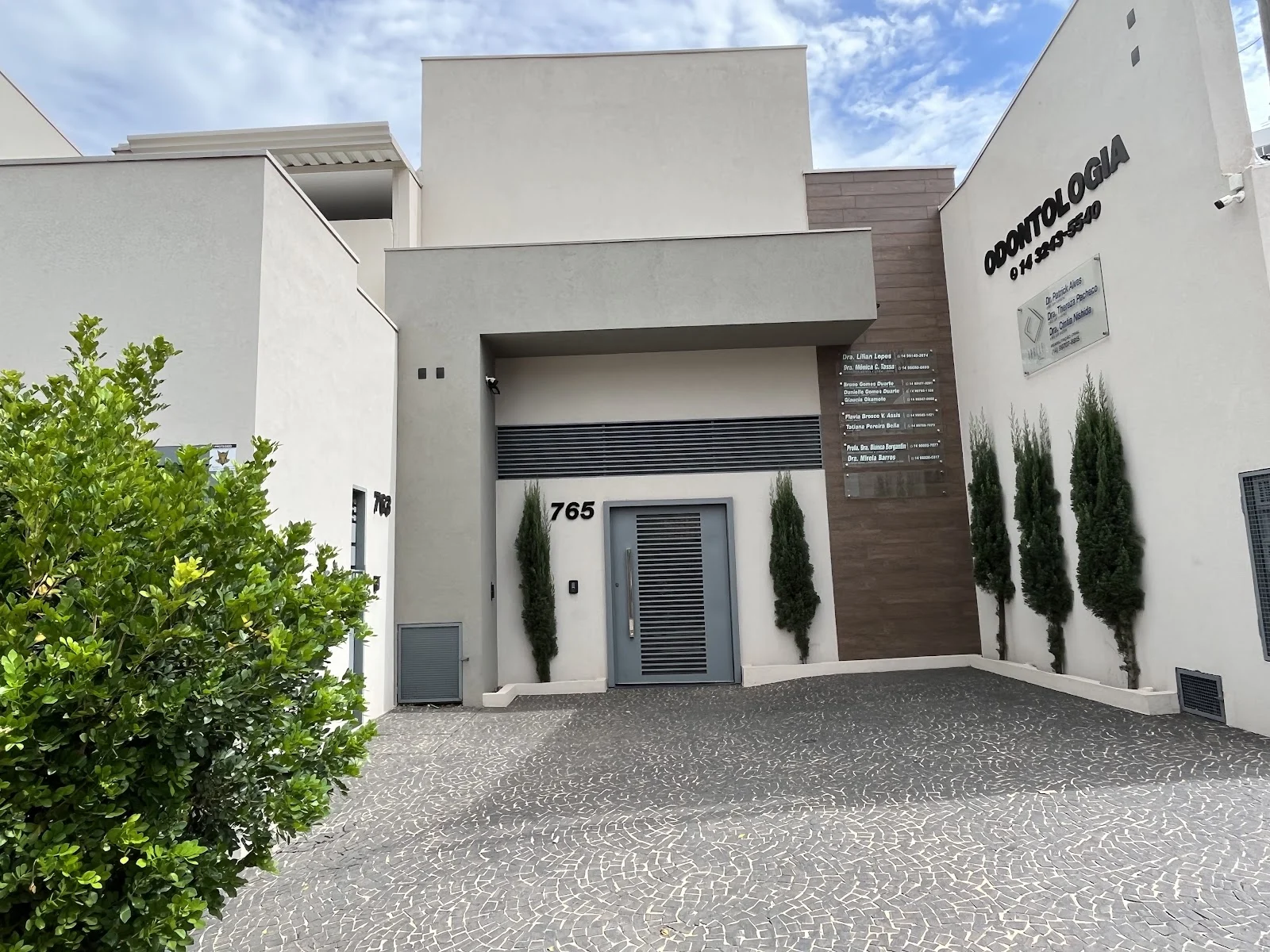 Fachada do prédio de consultórios no número 765 da Rua Horton Hoover: porta cinza, ciprestes e, ao lado, o painel de placas dos dentistas, entre elas a da Dra. Mirela Barros.