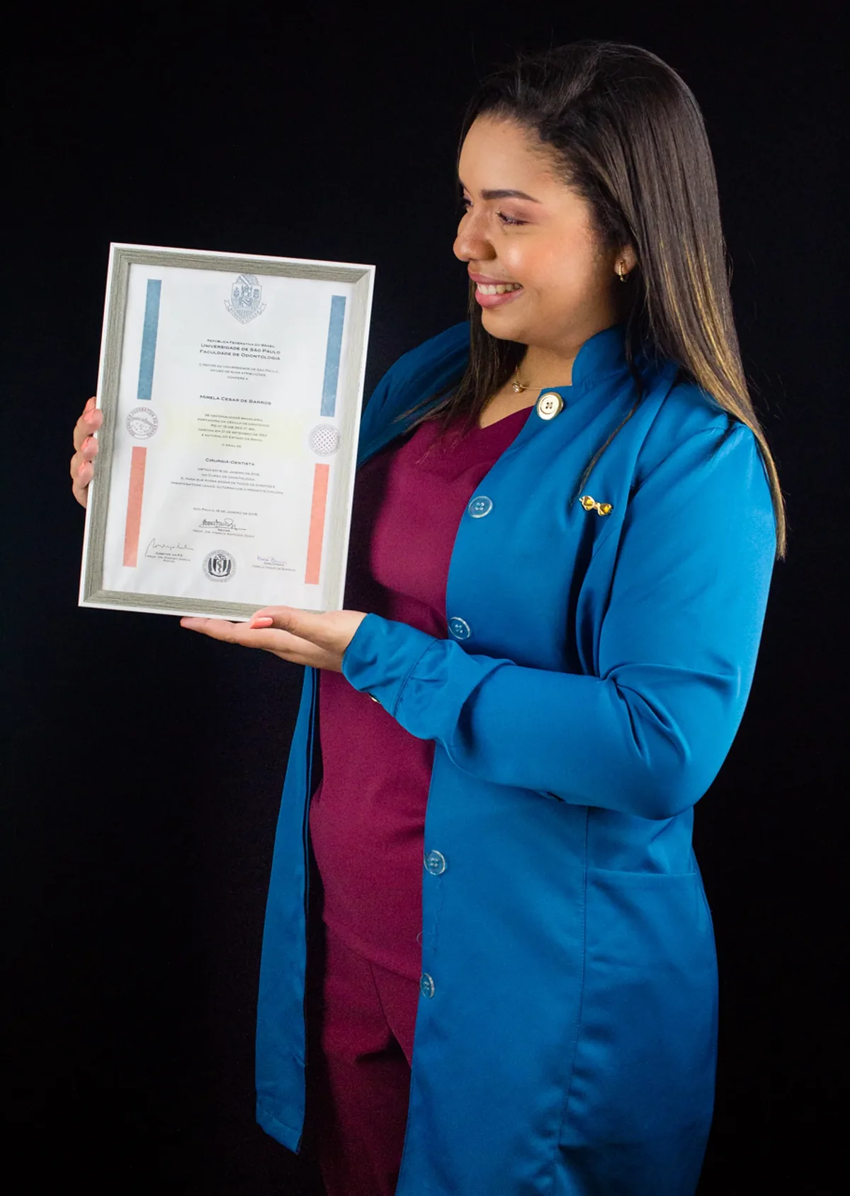 A Dra. Mirela Barros, de jaleco azul sobre scrub bordô, sorri olhando para o diploma de cirurgiã-dentista da Faculdade de Odontologia da USP, que segura emoldurado.