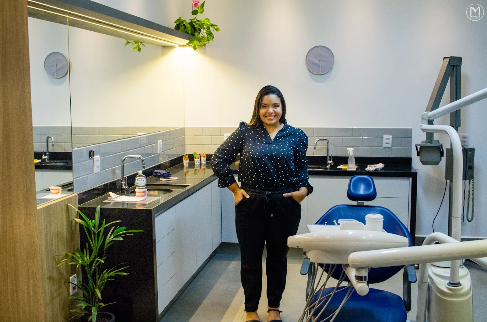 A Dra. Mirela Barros em pé no consultório, ao lado da cadeira odontológica azul-marinho.